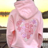 Pink hoodie with a decorative back design featuring shells and starfish, set against a blurred natural background.